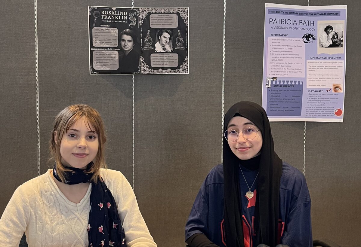 deux étudiants assis à une table avec des affiches de projets sur un mur derrière eux