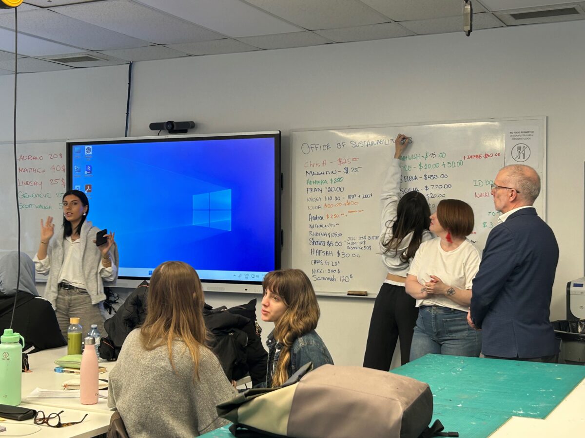 des élèves dans une salle de classe écoutent un présentateur pendant qu'un élève écrit les montants des dons sur un tableau blanc