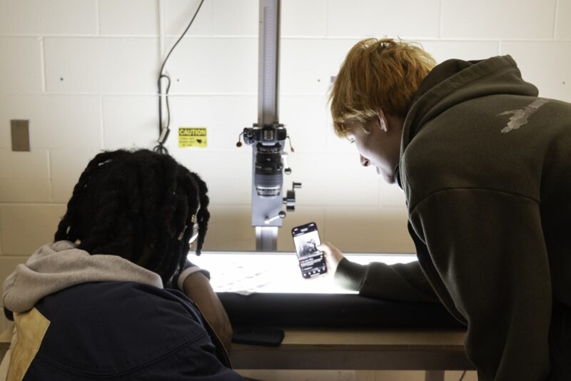 Deux étudiants en photographie discutent autour d'une table lumineuse et regardent un clip vidéo sur un téléphone portable.